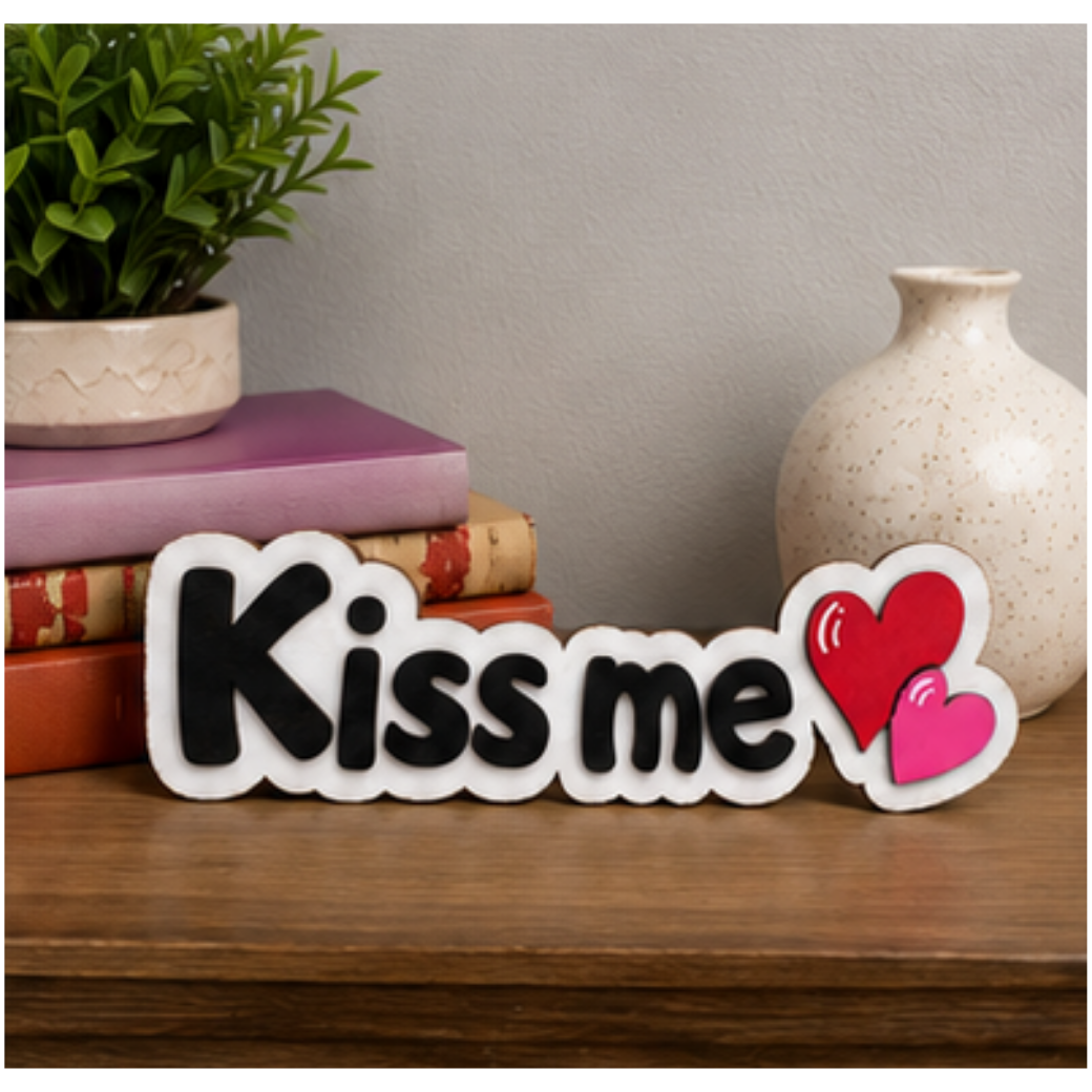 Kiss Me layered wood shelf sitter displayed on a wooden table beside decorative books, a potted plant, and a ceramic vase in a cozy home setting.
